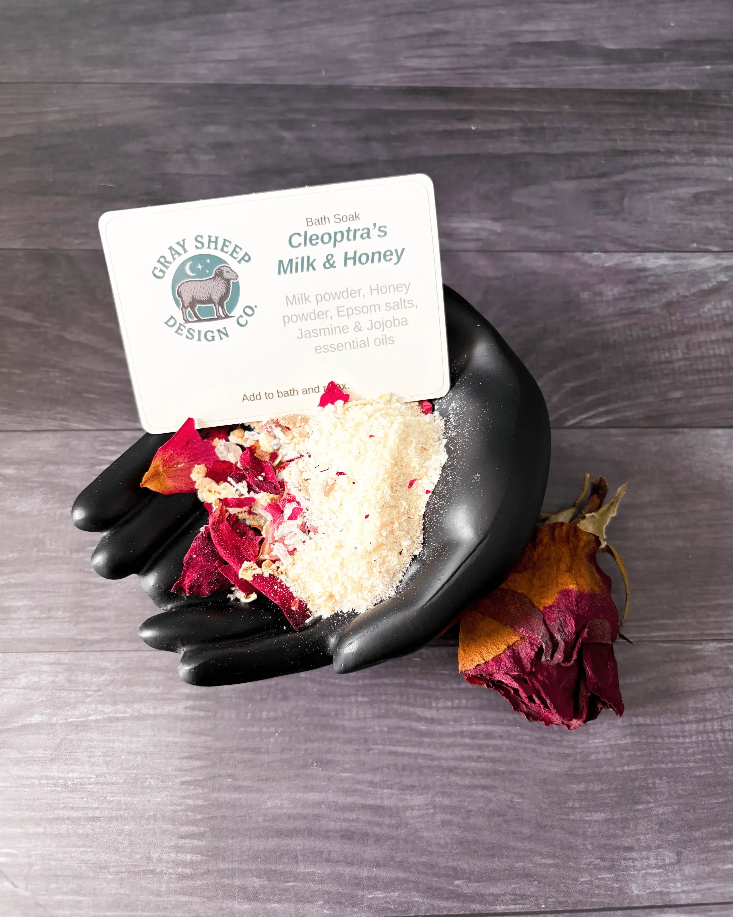 Loose Cleopatra's Milk & Honey soak in a black hand-shaped dish — creamy powder with rose petals and a dried rose.