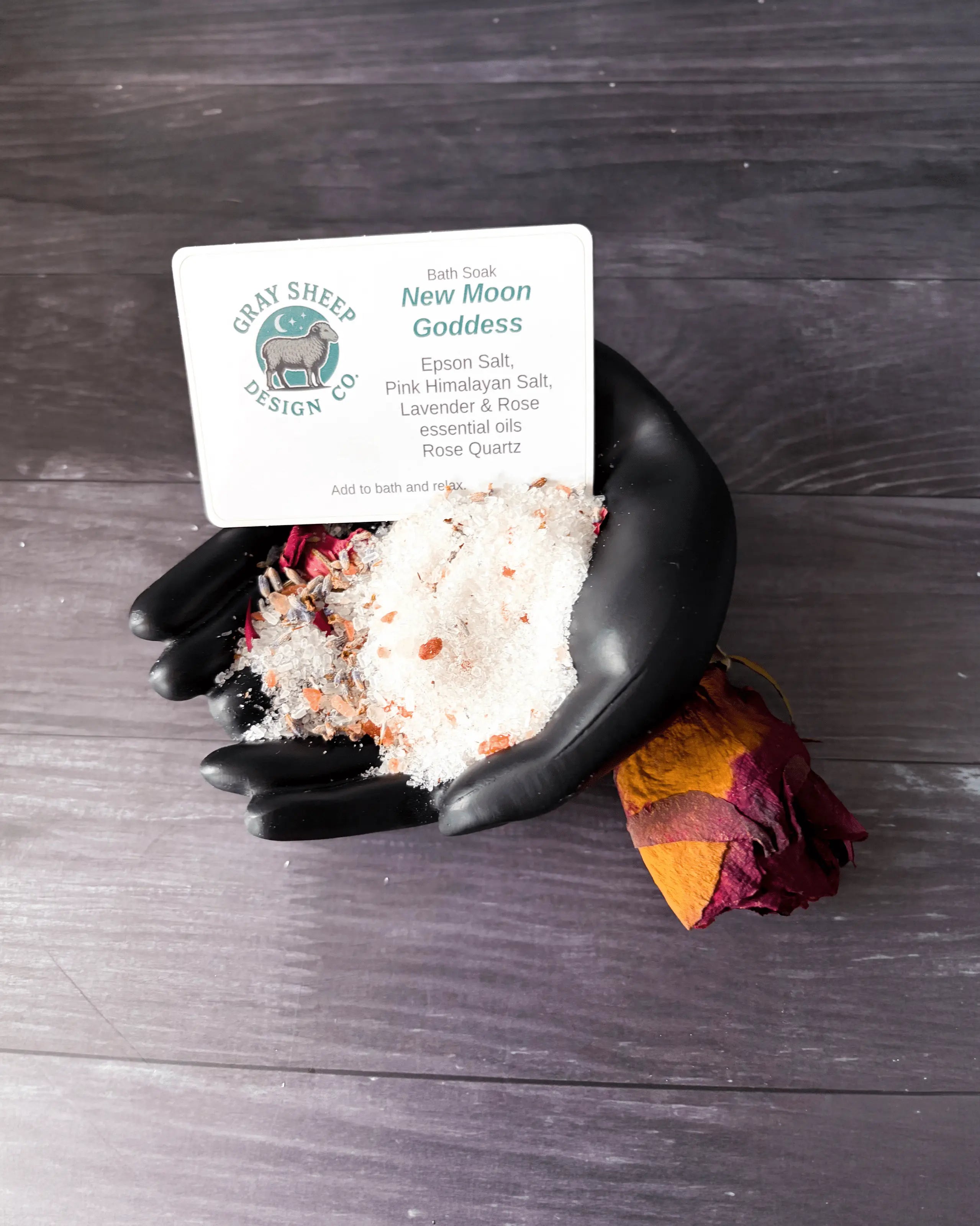 Loose New Moon Goddess soak in a black hand-shaped dish — pink-and-white salts with a dried rose and rose quartz.