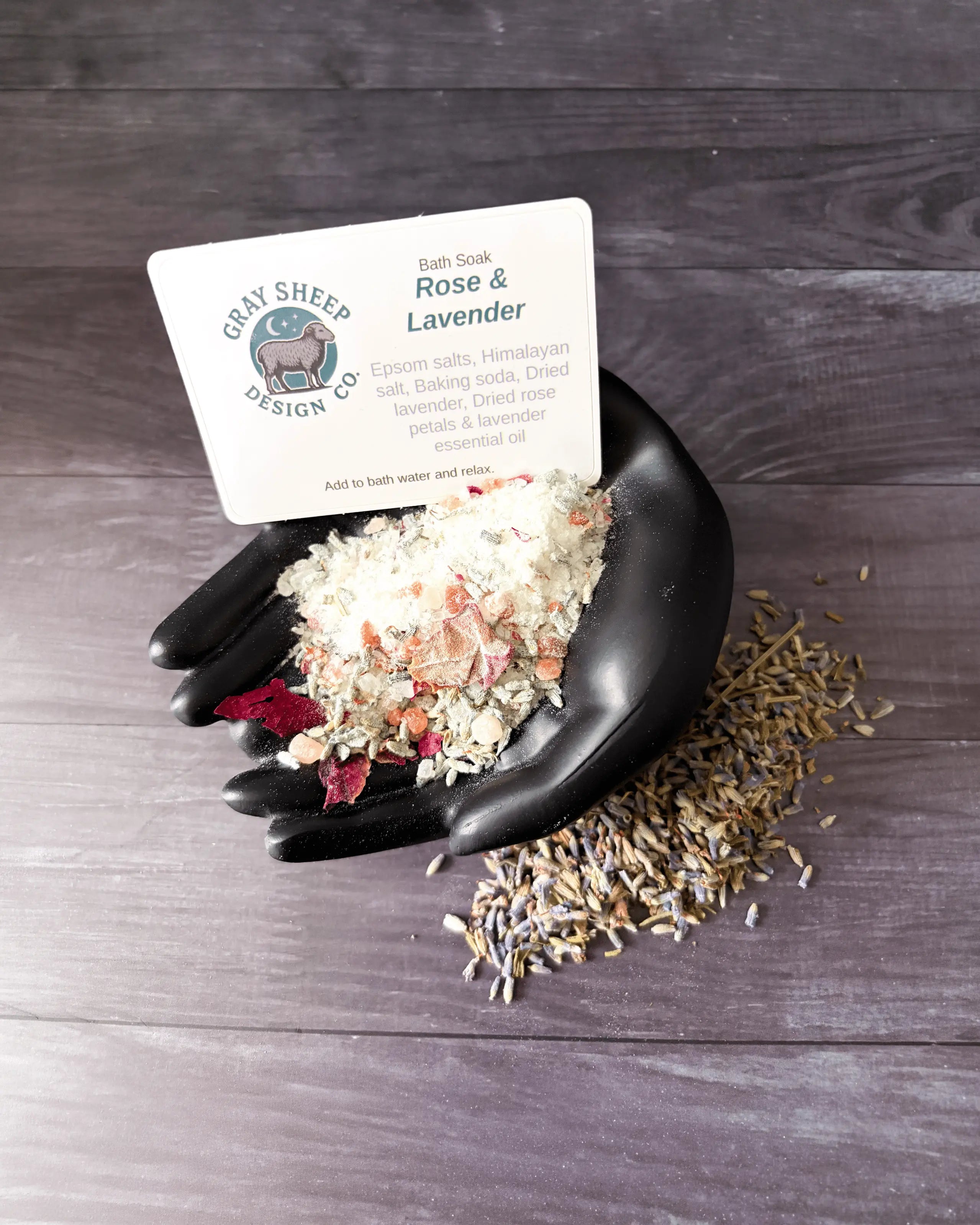 Loose Rose & Lavender soak in a black hand-shaped dish — pale salts, rose petals, and dried lavender.