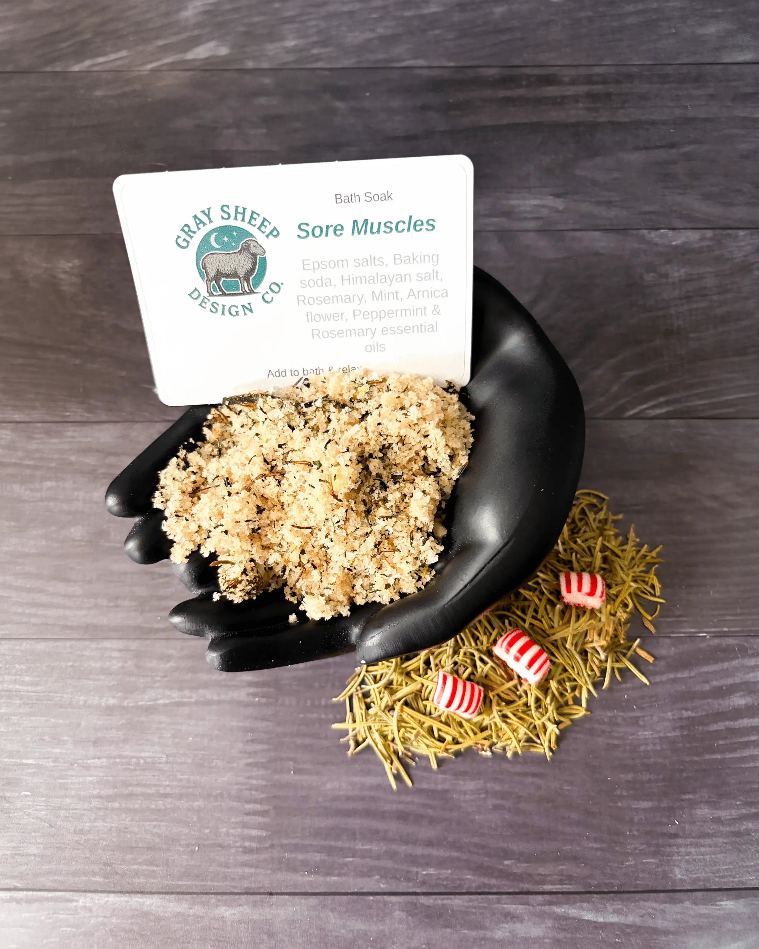 Loose Sore Muscles soak in a black hand-shaped dish — herbal bath salts with peppermint pieces and dried arnica.