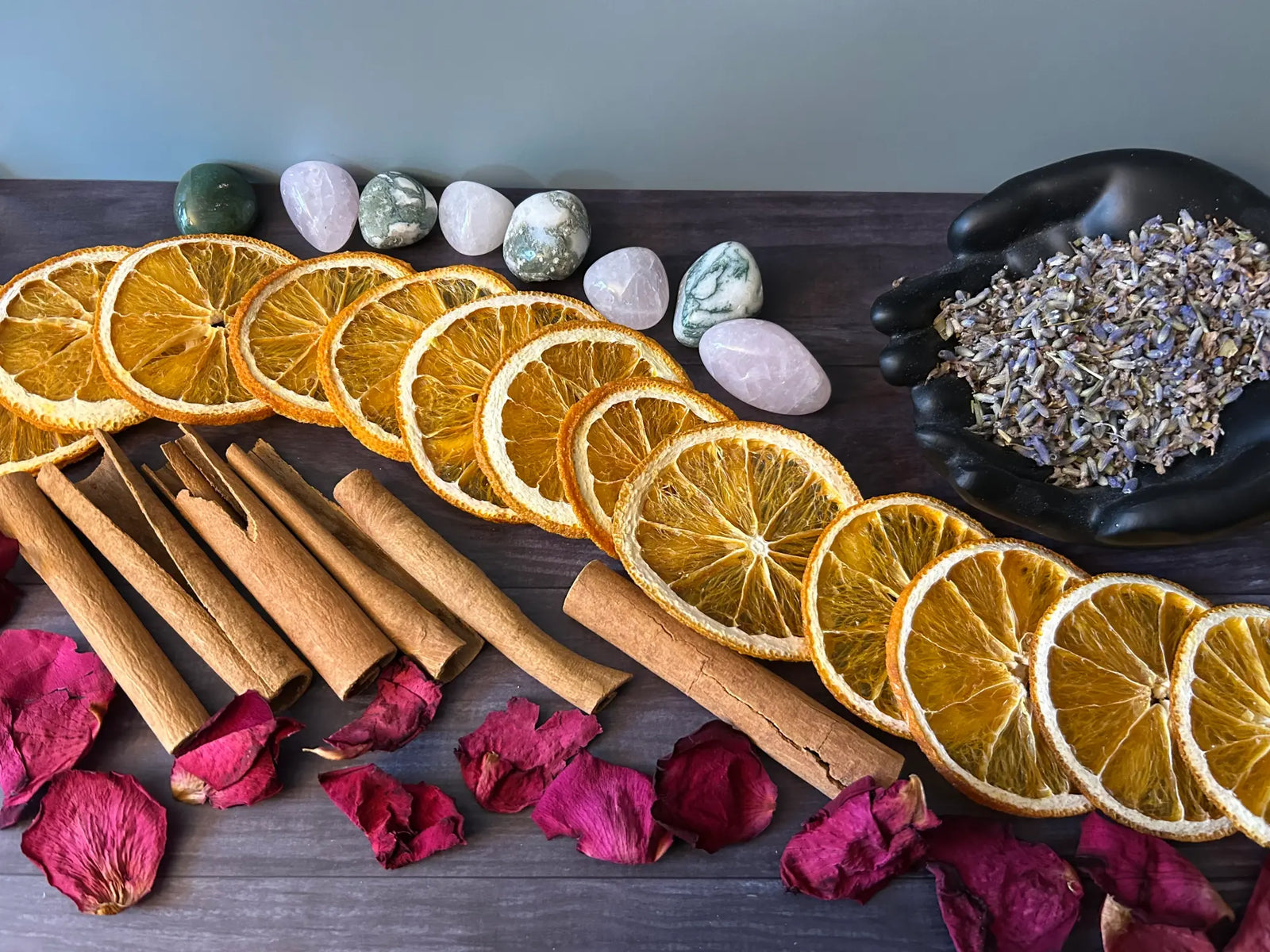 Dried orange slices, cinnamon sticks, rose petals, lavender, and polished stones arranged on a dark wood table.
