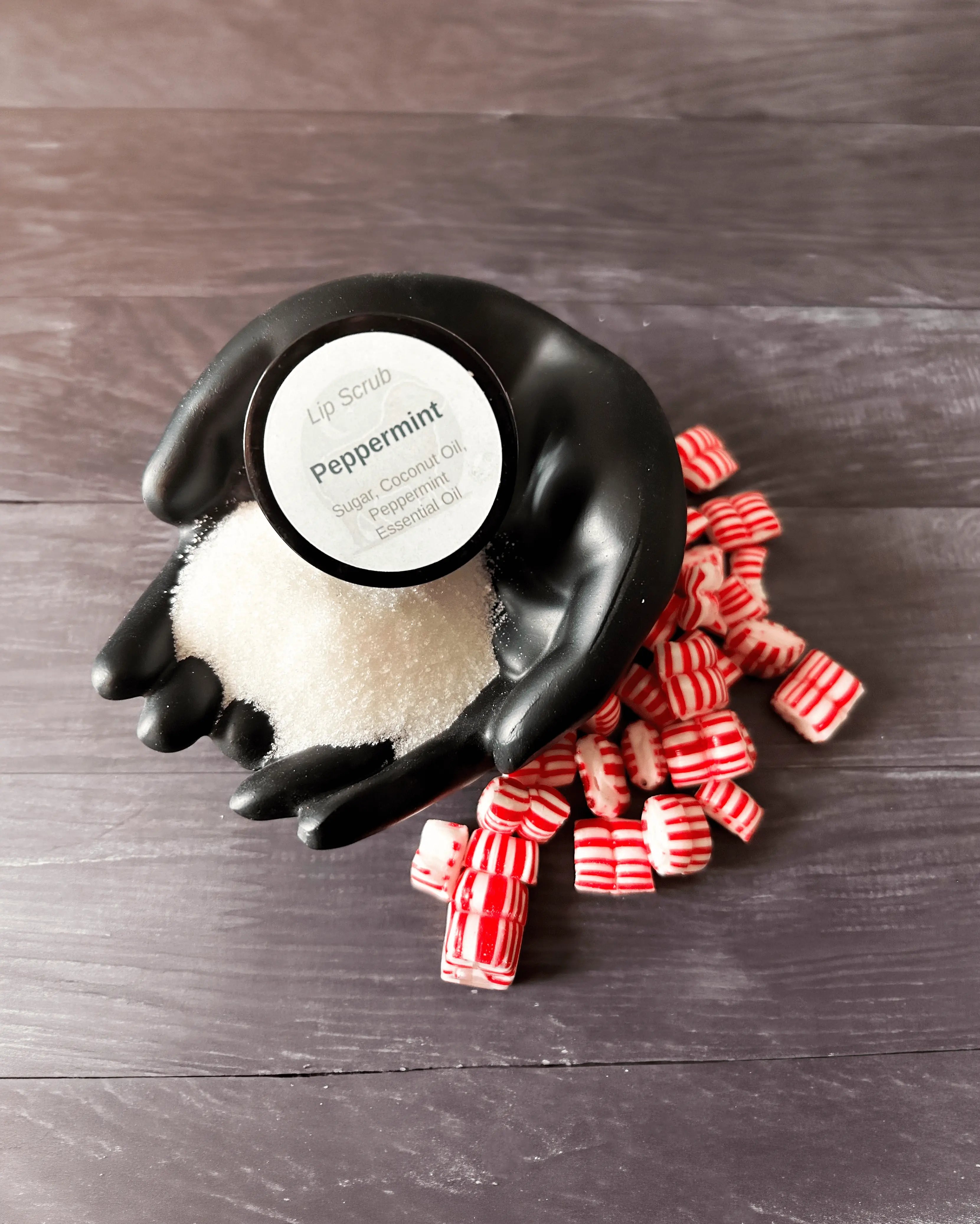 Open jar of Peppermint lip scrub in a black hand-shaped dish, surrounded by striped peppermint candies.