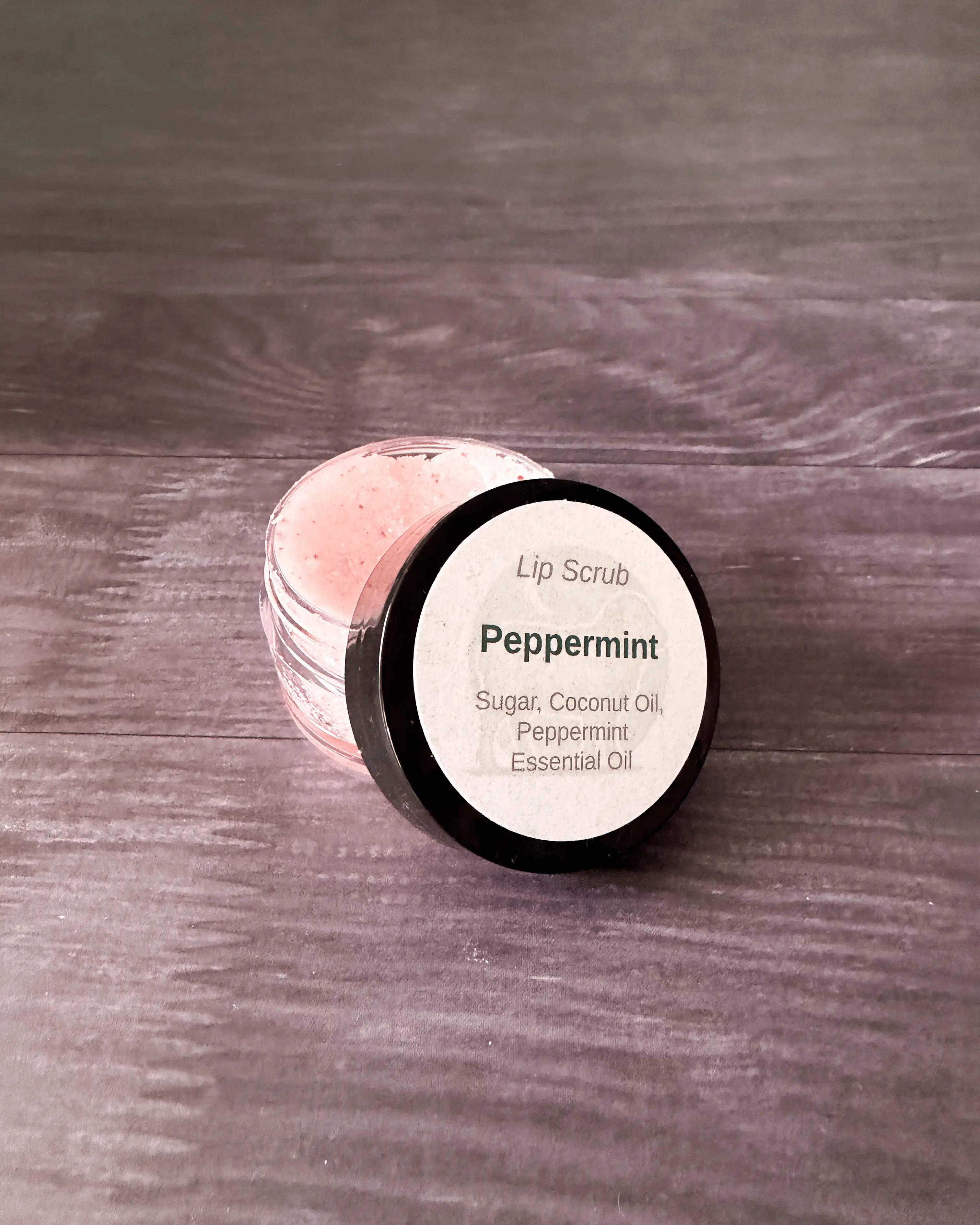 Peppermint lip scrub in a small clear jar with a black lid, pink-flecked sugar scrub inside.