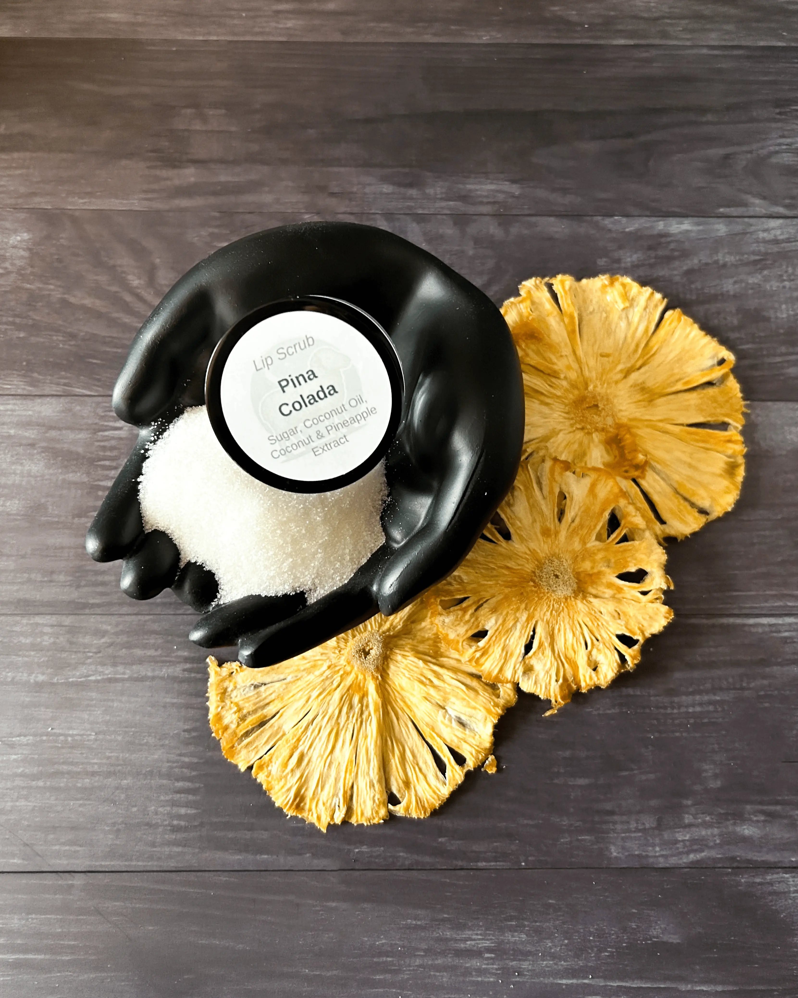 Open jar of Piña Colada lip scrub in a black hand-shaped dish with dried pineapple rings.