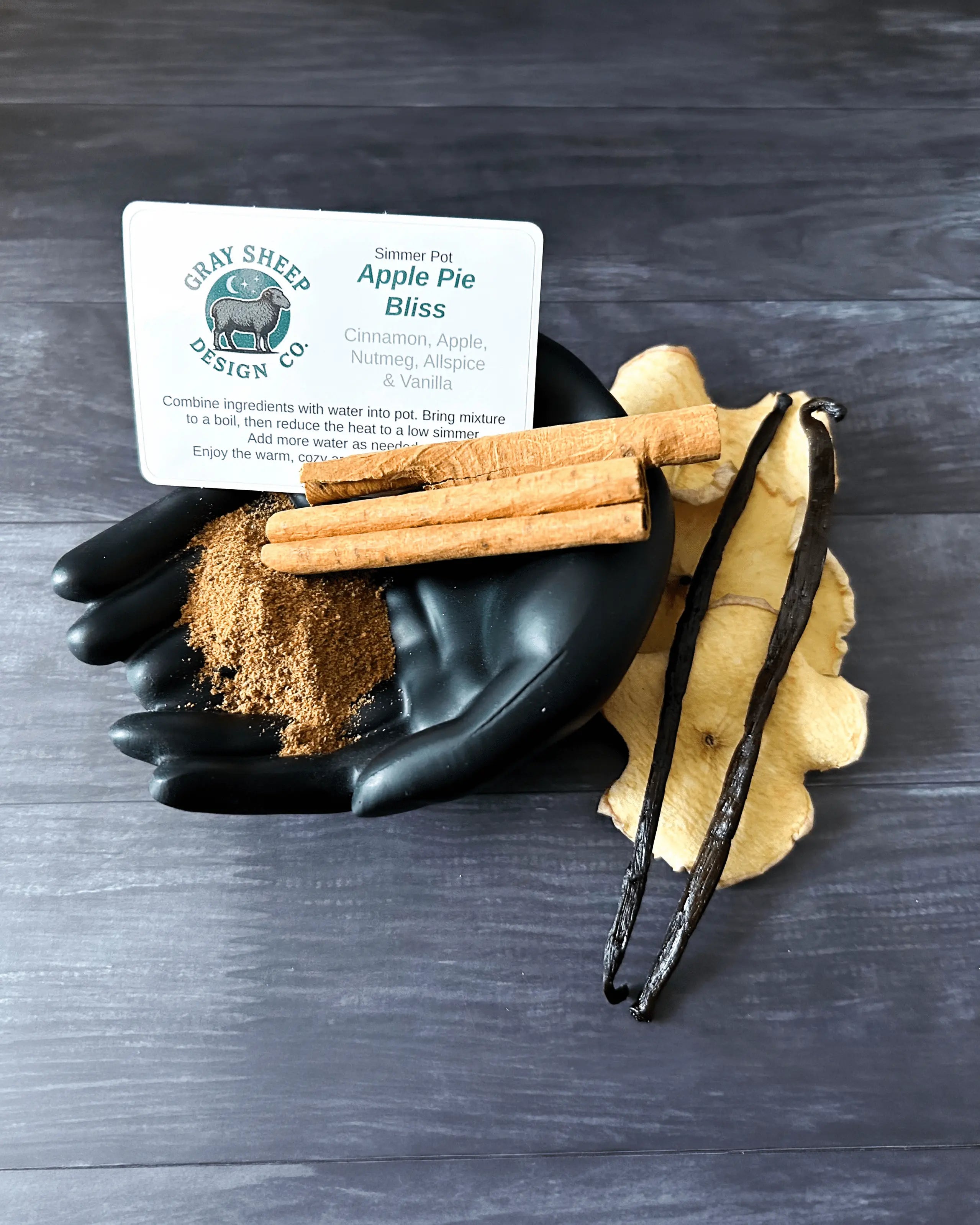 Loose Apple Pie Bliss blend in a black hand-shaped dish — cinnamon sticks, vanilla beans, dried apples, and ground spice.