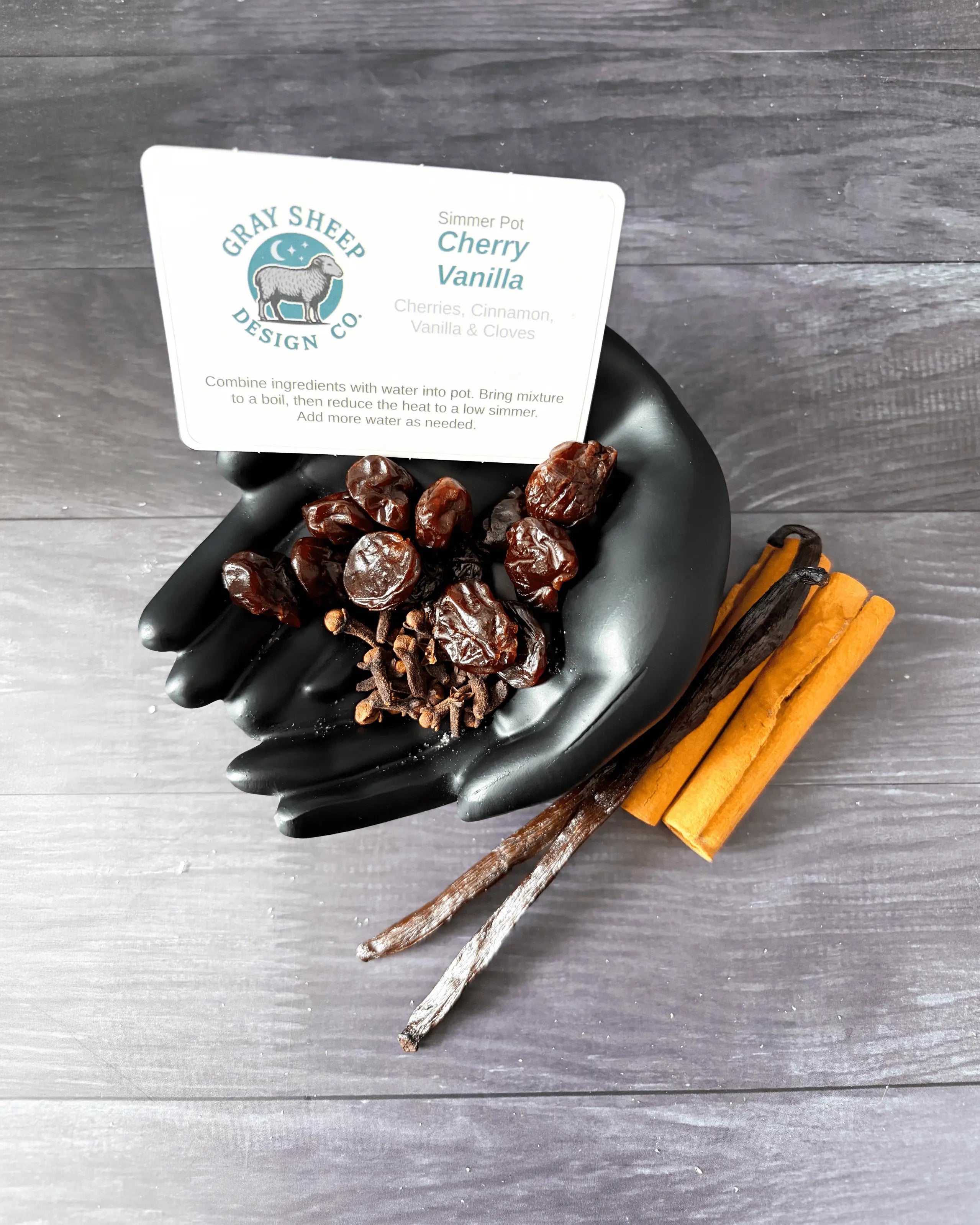 Loose Cherry Vanilla blend in a black hand-shaped dish — dried cherries, cinnamon sticks, and vanilla beans.