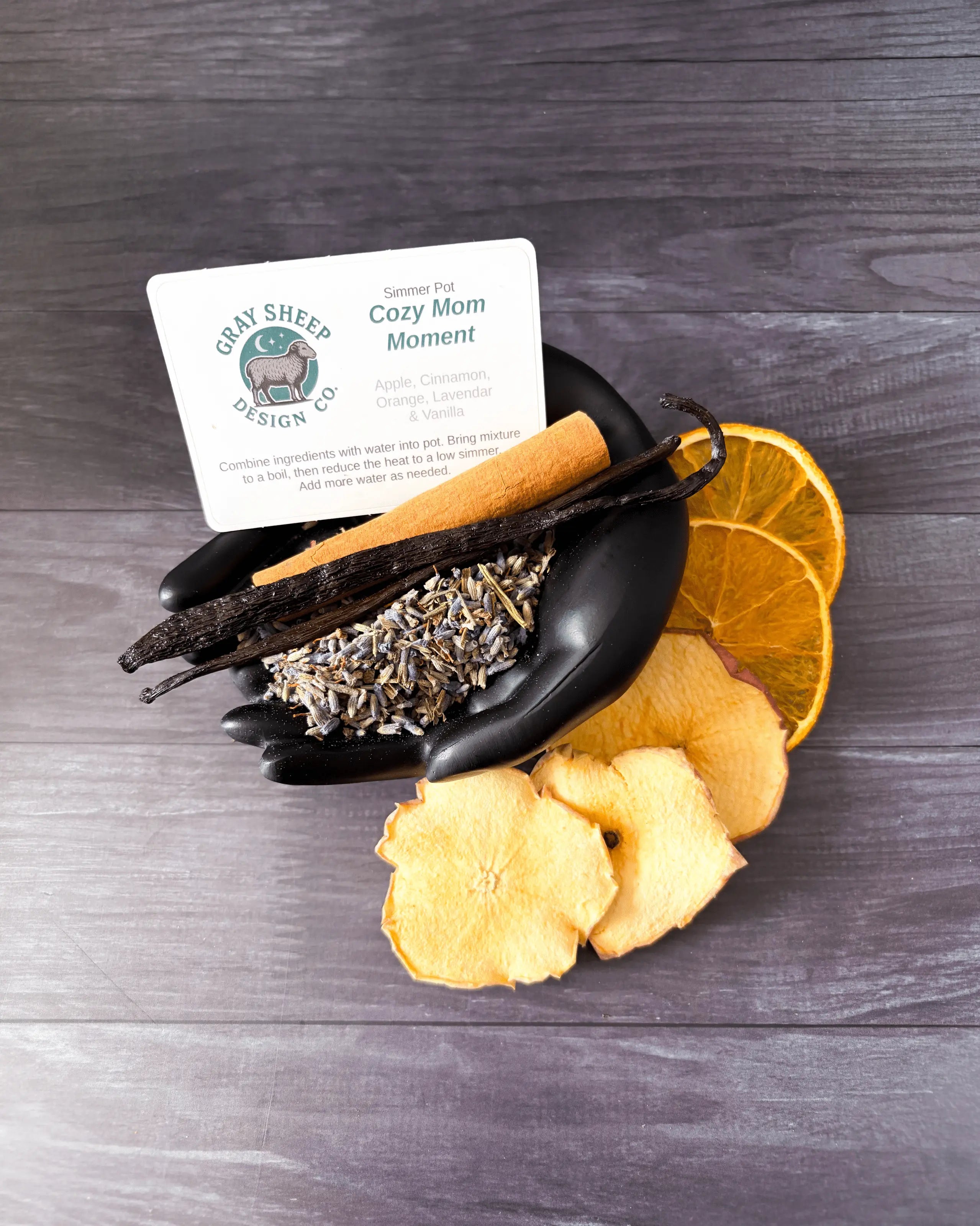 Loose Cozy Mom Moment blend in a black hand-shaped dish — lavender, vanilla beans, dried orange, and apple slices.