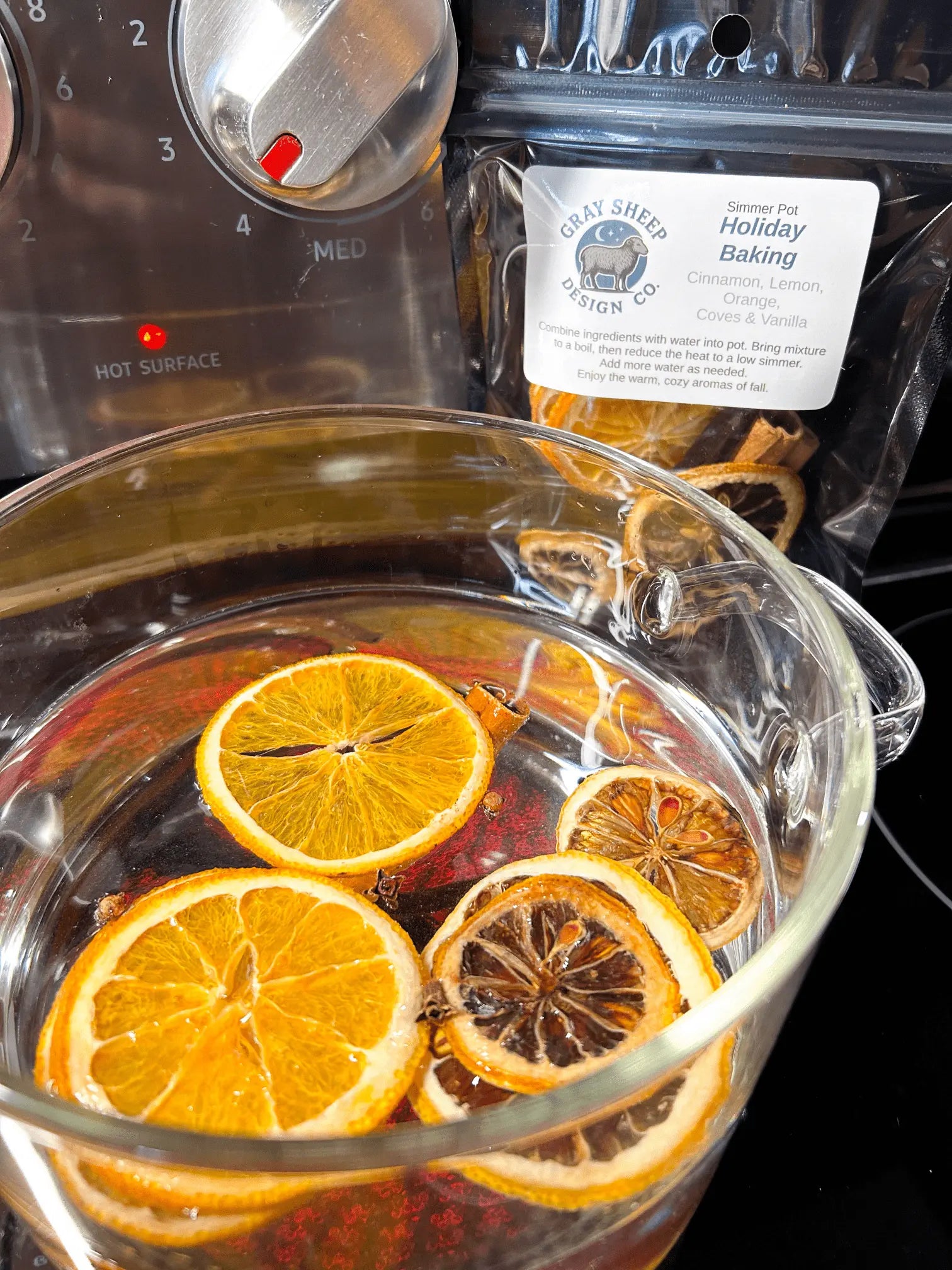 Holiday Baking simmer pot simmering on the stove — orange slices and spices in a glass pot, black pouch behind.